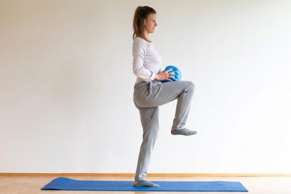 Die Trainerin steht aufrecht auf einem Bein, das andere ist angewinkelt, der Redondoball wird vor dem Bauch gehalten.